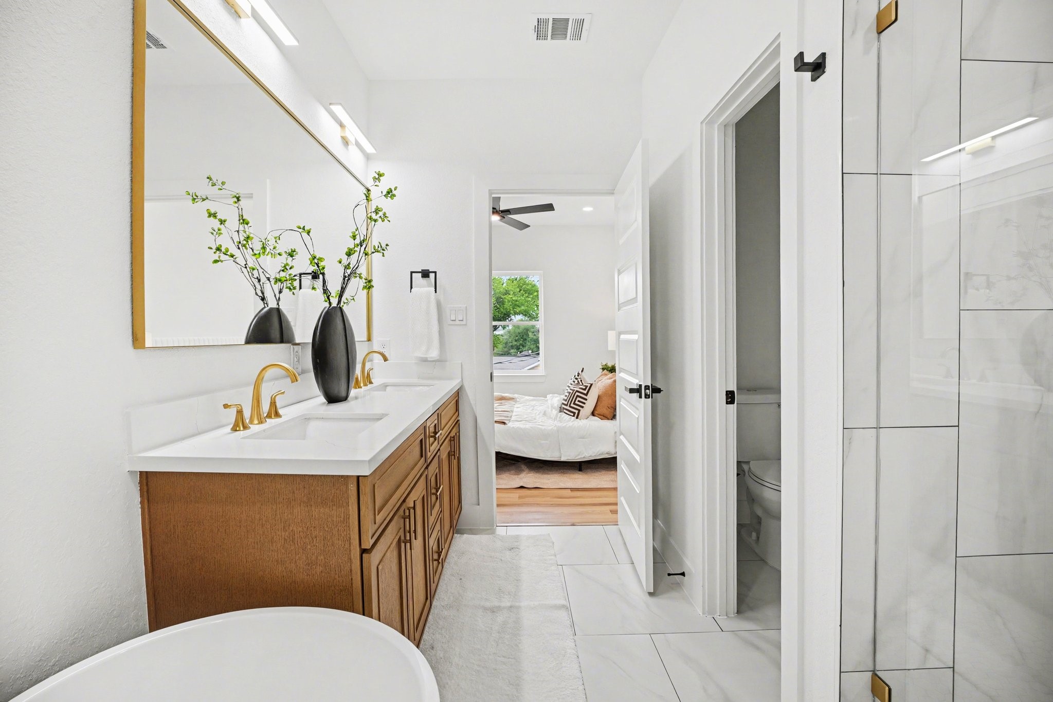115 Milby Street Houston, TX 77003 - Photo 16 of 28 This photo showcases a modern bathroom with a spacious double vanity featuring gold fixtures, a large mirror, and decorative vases. The room is bright with white and wood accents, and there's a partial view into a cozy bedroom with a large window, hardwood floors, and neutral decor.