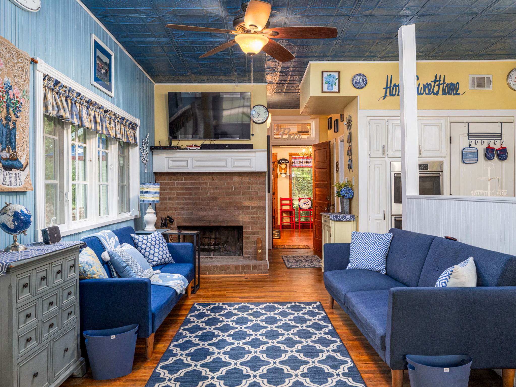 1065 Private Road 7041 Elgin, TX 78621 - Photo 7 of 38 Living room showcases stunning architectural details, including an ornate, vintage tin ceiling and rich oak wood floors. The space flows seamlessly to the additional kitchen area and office, enhanced by a charming brick fireplace and ceiling fan. Large windows fill the room with natural light, creating an inviting and cohesive living experience