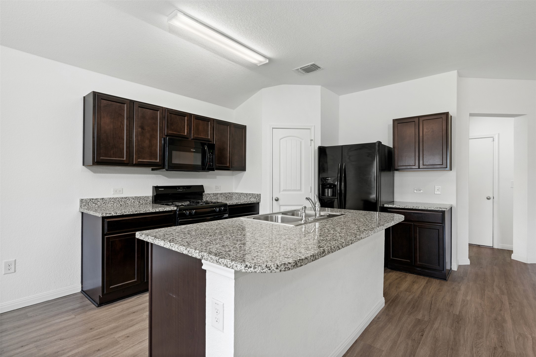 14900 Retha Pass Austin, TX 78725 - Photo 10 of 39 Kitchen featuring an island with a dual basin stainless steel sink, dark wood-finish cabinetry, granite countertops, and wood-finish flooring