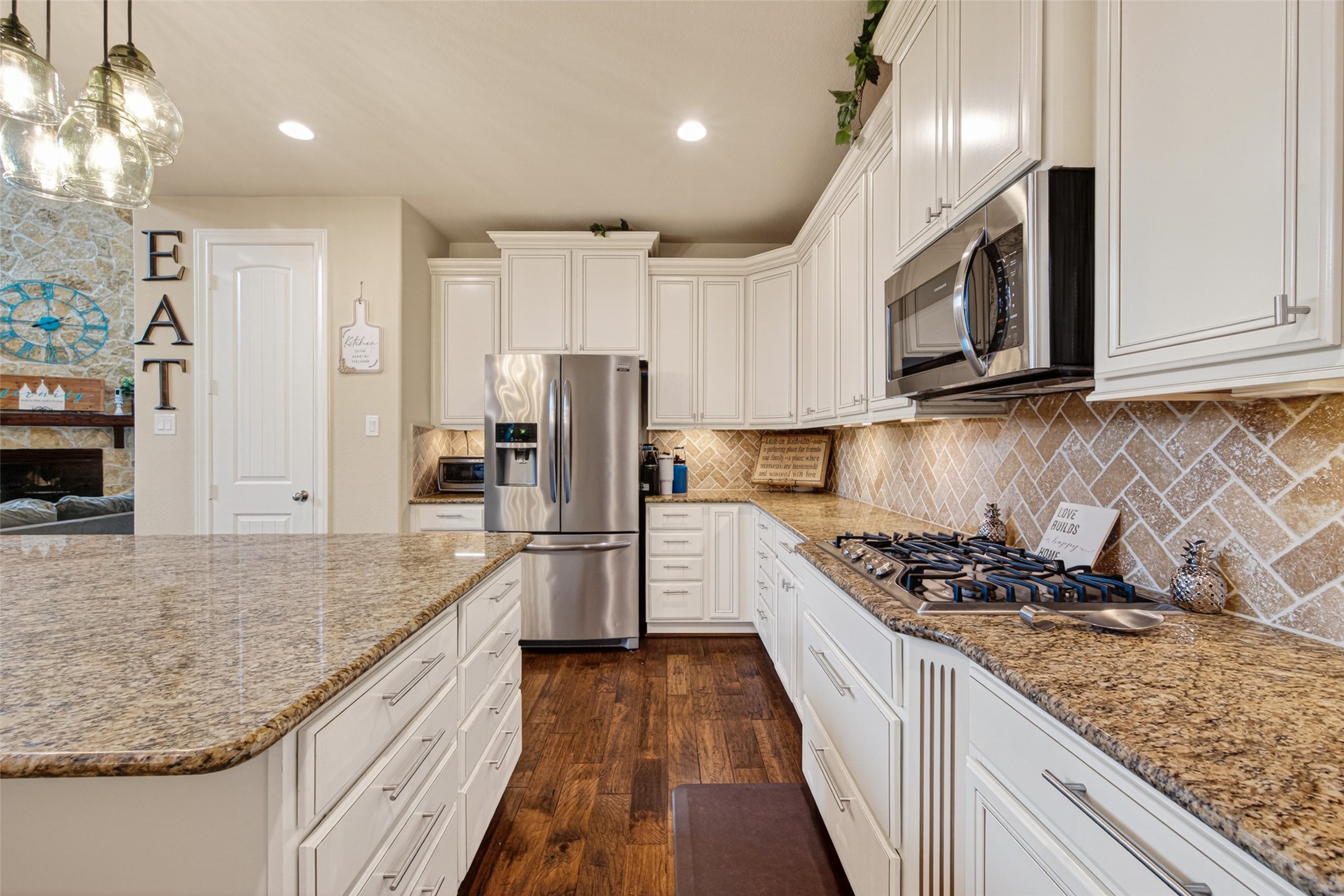 25611 Ellis Ridge Court Katy, TX 77494 - Photo 15 of 49 This kitchen features elegant white cabinetry, granite countertops, and stainless steel appliances. It includes a spacious island, a gas cooktop, and a warm, inviting backsplash. The open layout extends to a cozy living area with a stone fireplace.