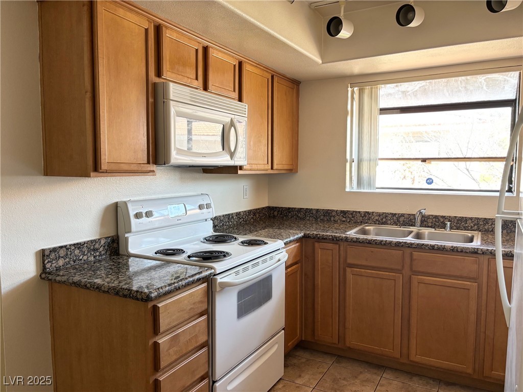 7300 West Pirates Cove Road, Unit 2054 Las Vegas, NV 89145 - Photo 5 of 16 Kitchen featuring white appliances, brown cabinetry, dark countertops, a sink, and light tile patterned flooring