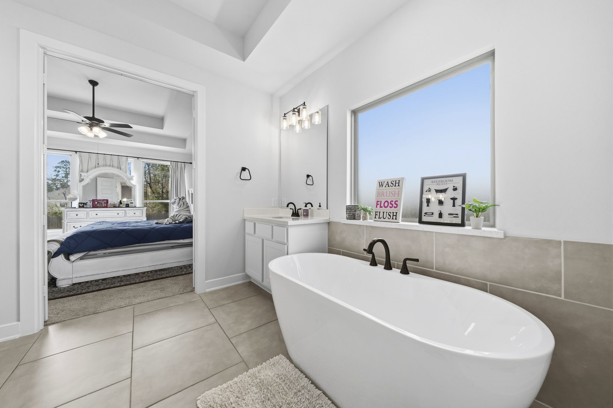 511 Carina Gaze Drive Conroe, TX 77318 - Photo 18 of 45 Bright, modern bathroom with a freestanding tub, large window for natural light, and sleek fixtures. It connects to a cozy bedroom with a ceiling fan and elegant décor. Perfect for relaxation and comfort.