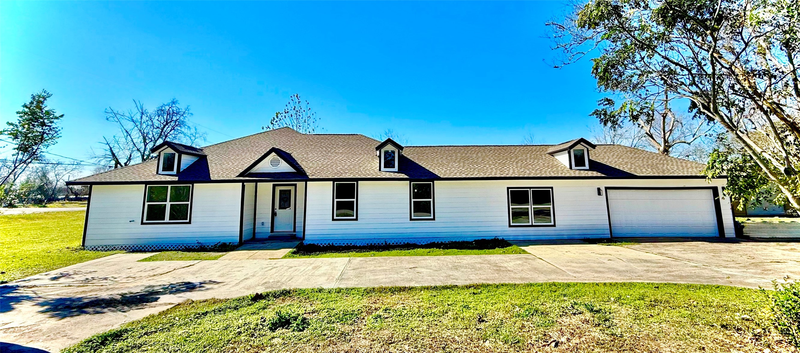 This photo shows a single-story, modern farmhouse-style home with a white exterior and dark roof. It features three dormer windows, a two-car garage, and a spacious driveway. The house is set on a large, grassy lot with some surrounding trees, creating a serene and inviting atmosphere.