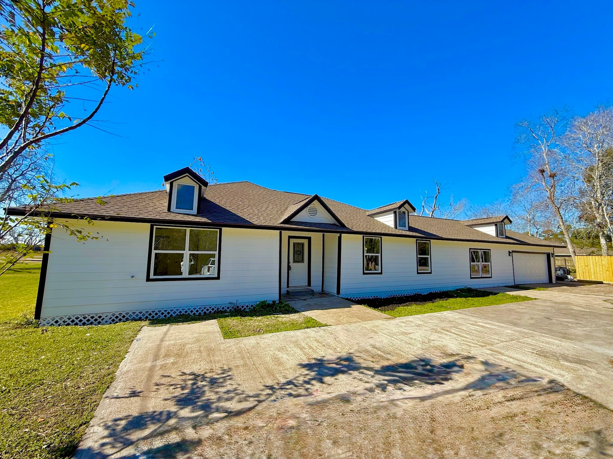 1214 Main Street Alvin, TX 77511 - Photo 2 of 18 Charming single-story home with a fresh white exterior and contrasting dark trim. It features dormer windows, a spacious driveway, and a two-car garage. The surrounding yard is open with a few trees, offering ample outdoor space.