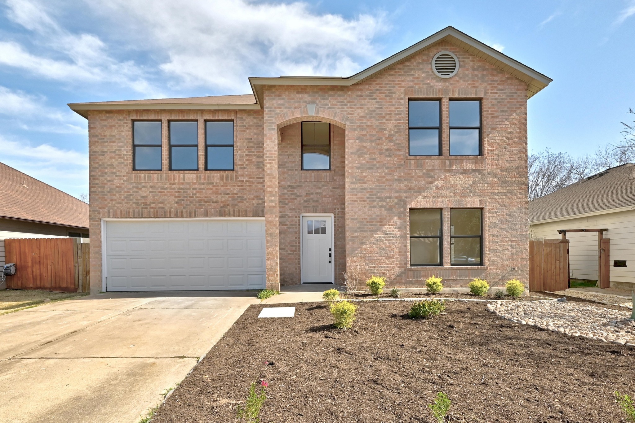 Two-story brick facade residence featuring a two-car garage, arched entryway, multiple windows with dark framing, and a newly prepared front yard with landscaping
