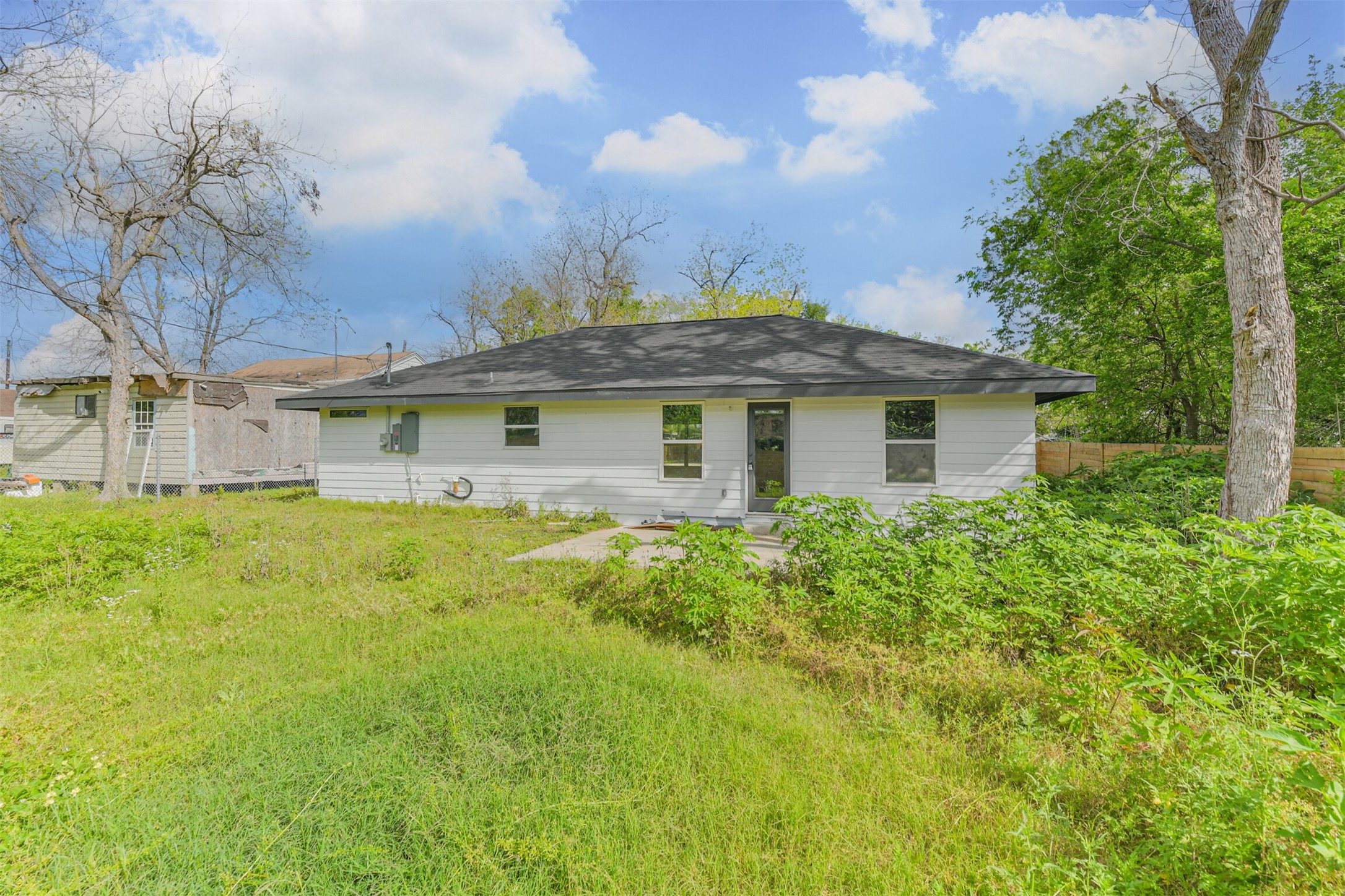 209 Leago Street Houston, TX 77022 - Photo 19 of 24 This photo showcases a single-story home with a modern, light-colored exterior and a dark roof. It sits on a spacious lot with a grassy yard and mature trees, offering potential for landscaping or gardening. The setting is peaceful, with neighboring homes visible in the background.