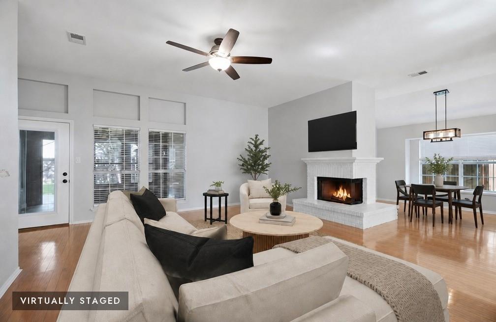 2074 Briarcliff Road Lewisville, TX 75067 - Photo 3 of 40 Spacious living area featuring a prominent white brick fireplace, wood-finish flooring, a ceiling fan, and multiple windows - virtually staged