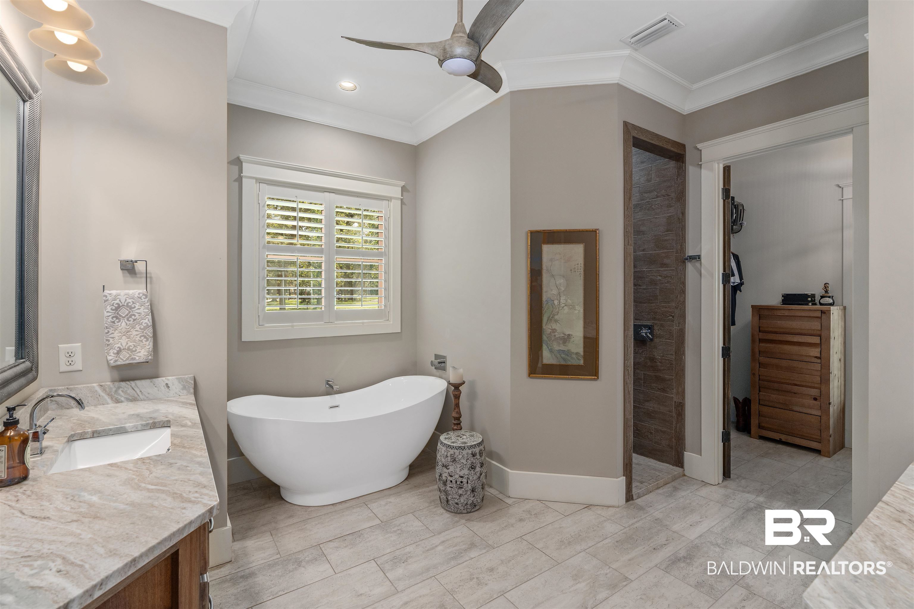 8753 Bishop Road Fairhope, AL 36532 - Photo 30 of 59 Freestanding soaking tub with a window featuring white plantation shutters, dual vanities with stone countertops, a walk-in shower with tiled walls, and wood-finish tile flooring