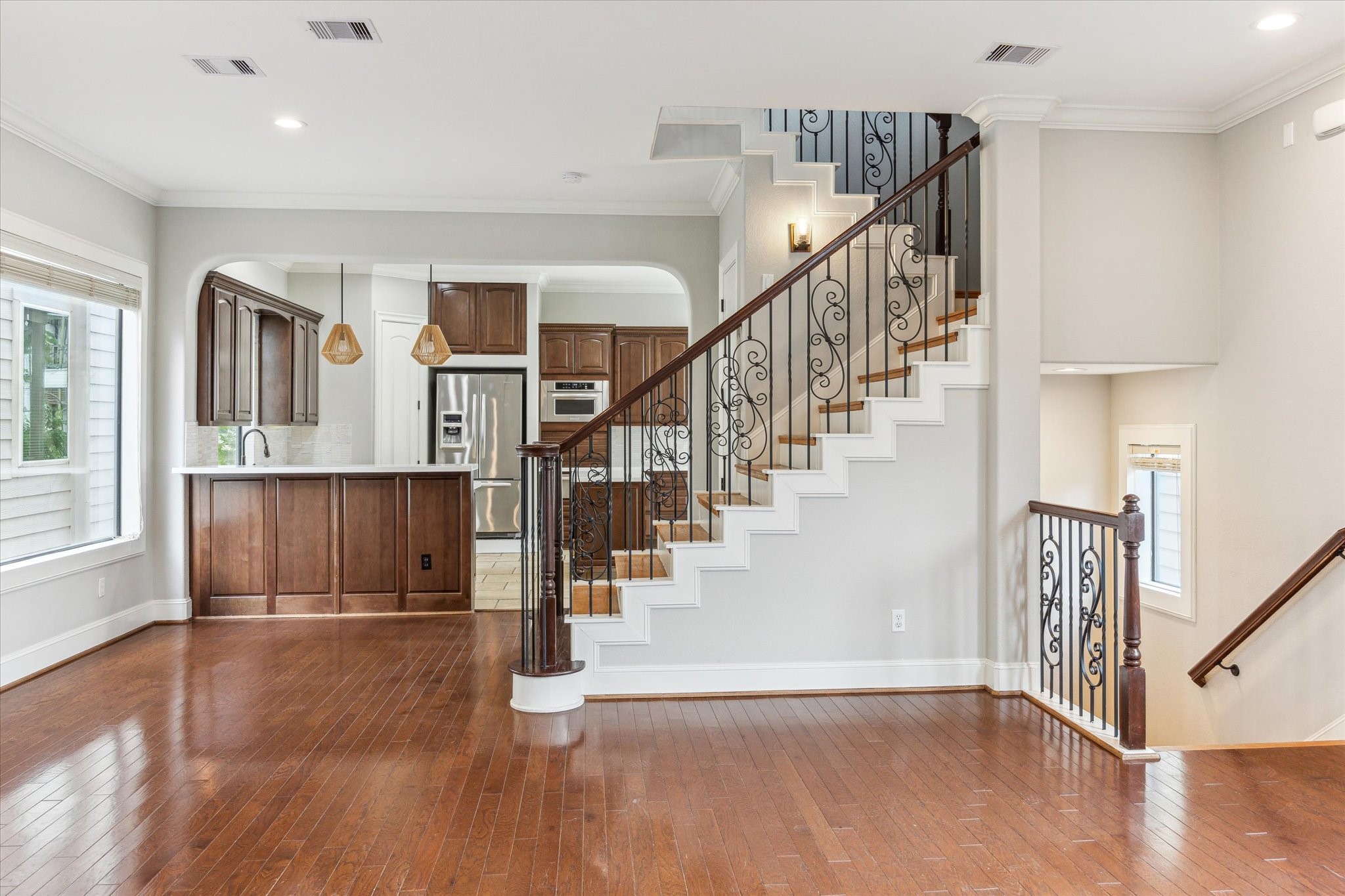 1512 Thompson Street, Unit 3 Houston, TX 77007 - Photo 12 of 32 This photo showcases a bright and open living space with hardwood floors, featuring a stylish staircase with wrought iron railings. The kitchen, visible from the living area, includes modern stainless steel appliances, rich wood cabinetry, and elegant pendant lighting. Large windows allow plenty of natural light to enhance the space.
