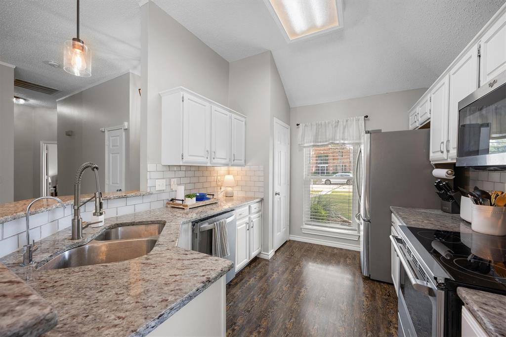 10714 Huntington Road Frisco, TX 75035 - Photo 4 of 24 Modern kitchen featuring white cabinetry, subway tile backsplash, dual stainless steel sinks with brushed nickel faucet, granite countertops, stainless steel appliances, wood-finish flooring, and abundant natural light.