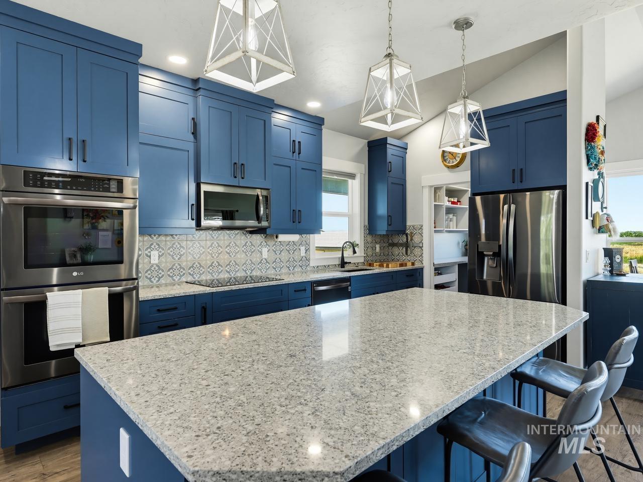 4950 West Columbia Road Nampa, ID 83687 - Photo 15 of 50 Spacious kitchen featuring an expansive island with a light-toned countertop, deep blue cabinetry, stainless steel appliances, a decorative tile backsplash, and contemporary pendant lighting