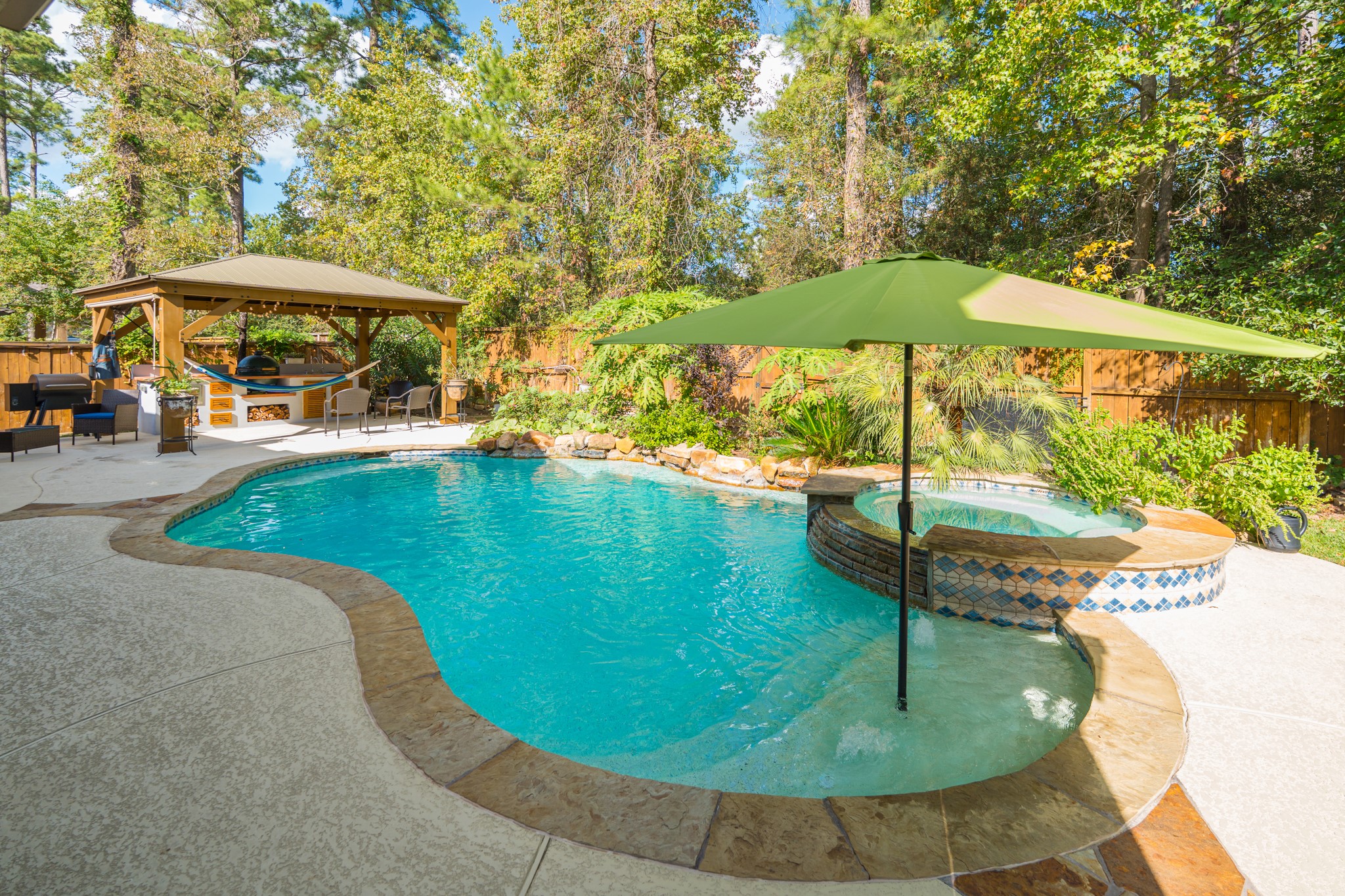 30 Fury Ranch Place The Woodlands, TX 77375 - Photo 44 of 47 This photo showcases a beautiful backyard with a sparkling pool and adjacent hot tub, surrounded by lush greenery. A shaded patio area with seating offers a perfect space for relaxation and entertaining.