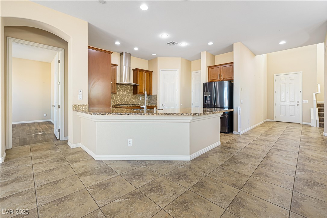 60 Newton Ridge Way Las Vegas, NV 89183 - Photo 3 of 44 Kitchen featuring decorative backsplash, brown cabinets, wall chimney range hood, light stone counters, and stainless steel fridge with ice dispenser