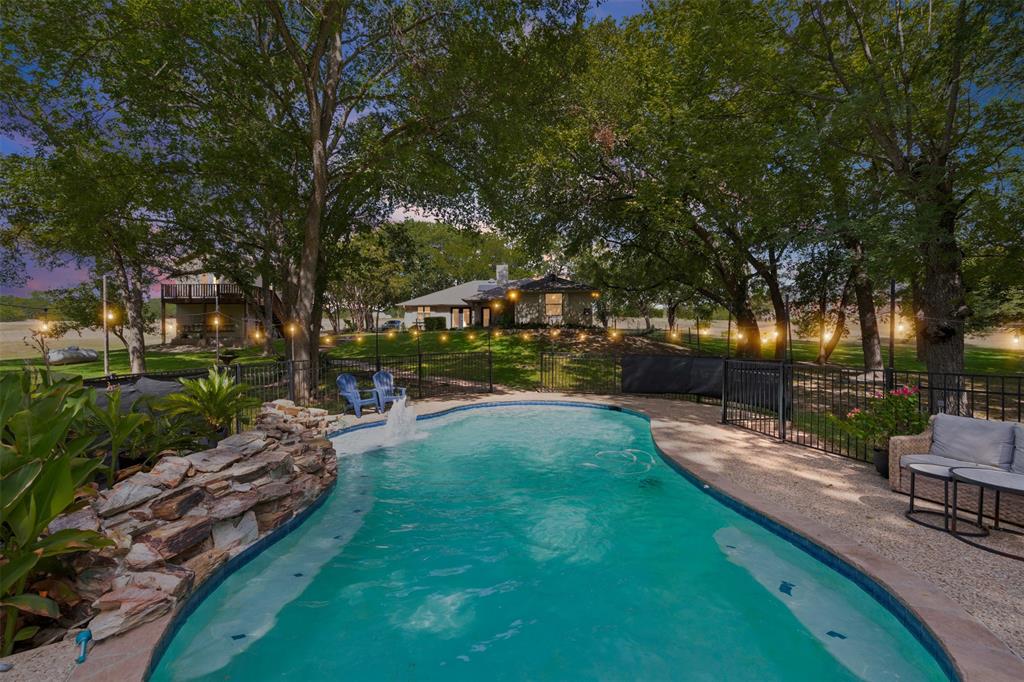 318 Ridge Creek Road Waxahachie, TX 75167 - Photo 1 of 40 View of property from back lot shows swimming pool with a rock waterfall feature, black metal perimeter fencing, and mature trees. Back of home and outbuilding with living space and balcony above can be seen in background