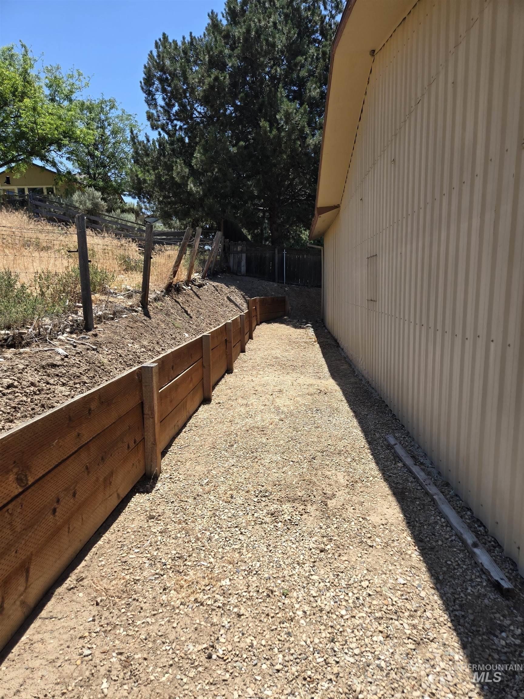 12050 West Hidden Valley Rim Road Boise, ID 83709 - Photo 32 of 39 Gravel pathway alongside a building with corrugated siding, bordered by a wooden retaining wall