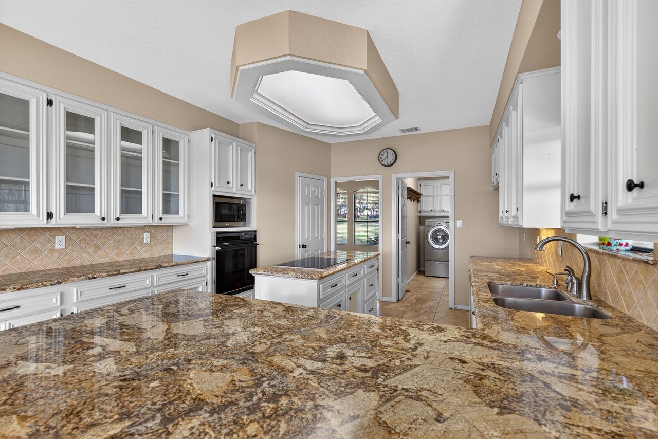 12626 Aries Loop Willis, TX 77318 - Photo 12 of 50 This kitchen features granite countertops, ample white cabinetry with glass-front doors, and modern appliances. It has a spacious layout with a central island and a view into a utility room with a washer and dryer. The neutral tile backsplash and floors complement the overall design.