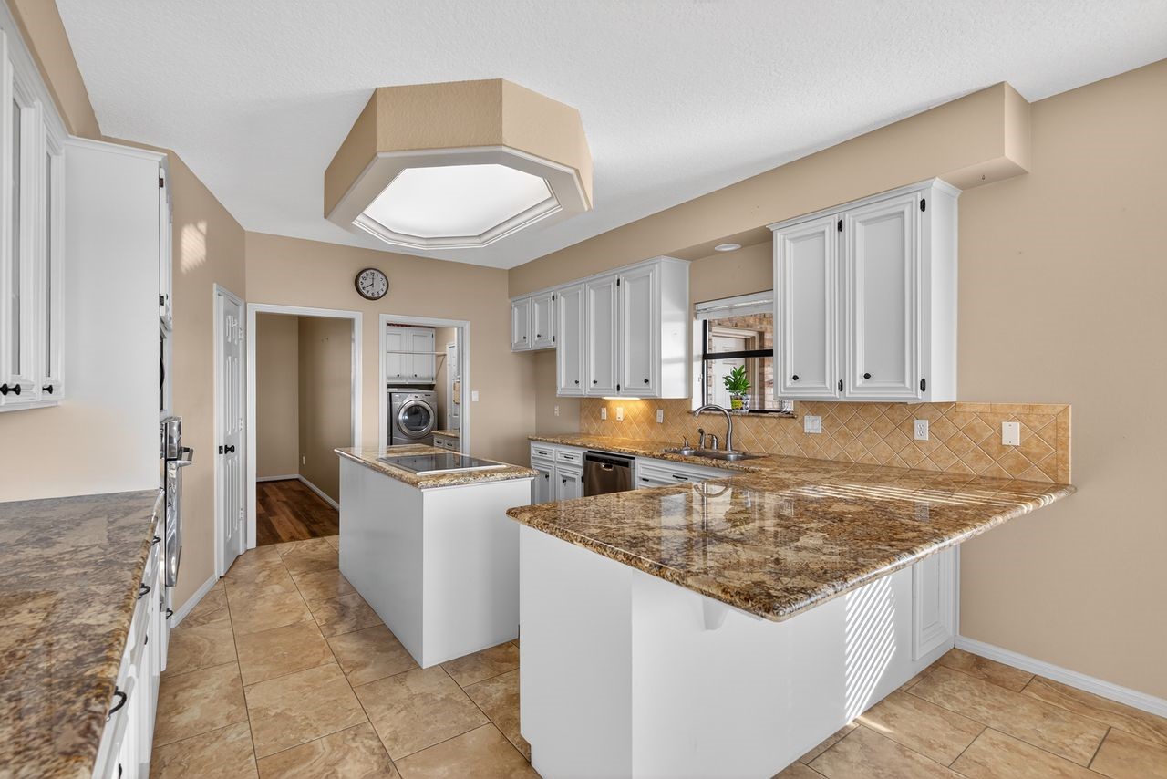 12626 Aries Loop Willis, TX 77318 - Photo 16 of 50 This kitchen features granite countertops, white cabinetry, and a central island. It has a built-in dishwasher, tiled backsplash, and an adjacent laundry area. The space is well-lit with natural light and overhead lighting, complemented by neutral-toned walls and floors.