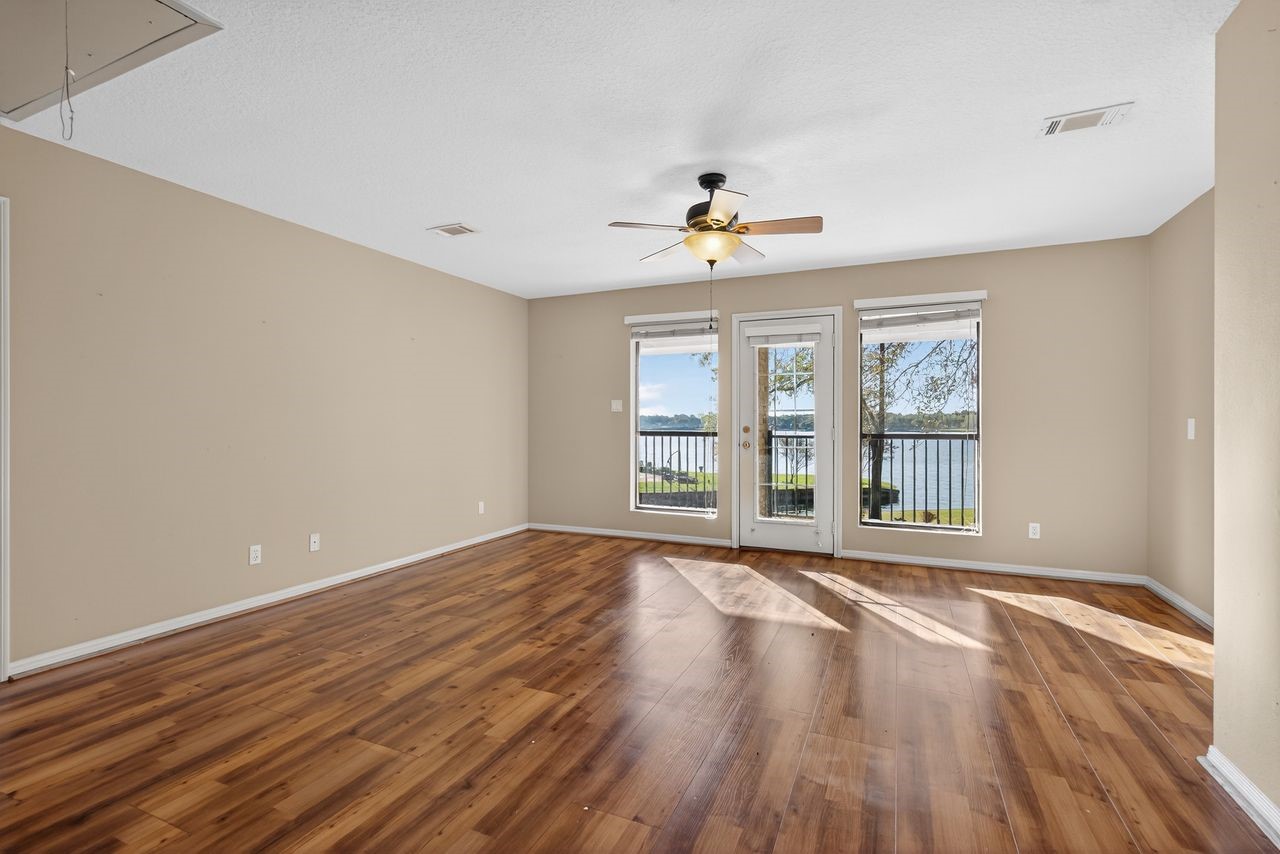 12626 Aries Loop Willis, TX 77318 - Photo 23 of 50 This spacious living room features rich wood flooring, neutral walls, and a ceiling fan. Large windows and a glass door offer abundant natural light and stunning lake views, creating a serene and inviting atmosphere.