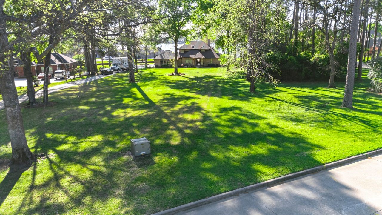 12626 Aries Loop Willis, TX 77318 - Photo 28 of 50 This photo showcases a spacious, grassy lot with mature trees providing shade, located in a serene residential neighborhood. Ideal for building a new home, the lot is surrounded by existing houses and has a clear, well-maintained lawn.