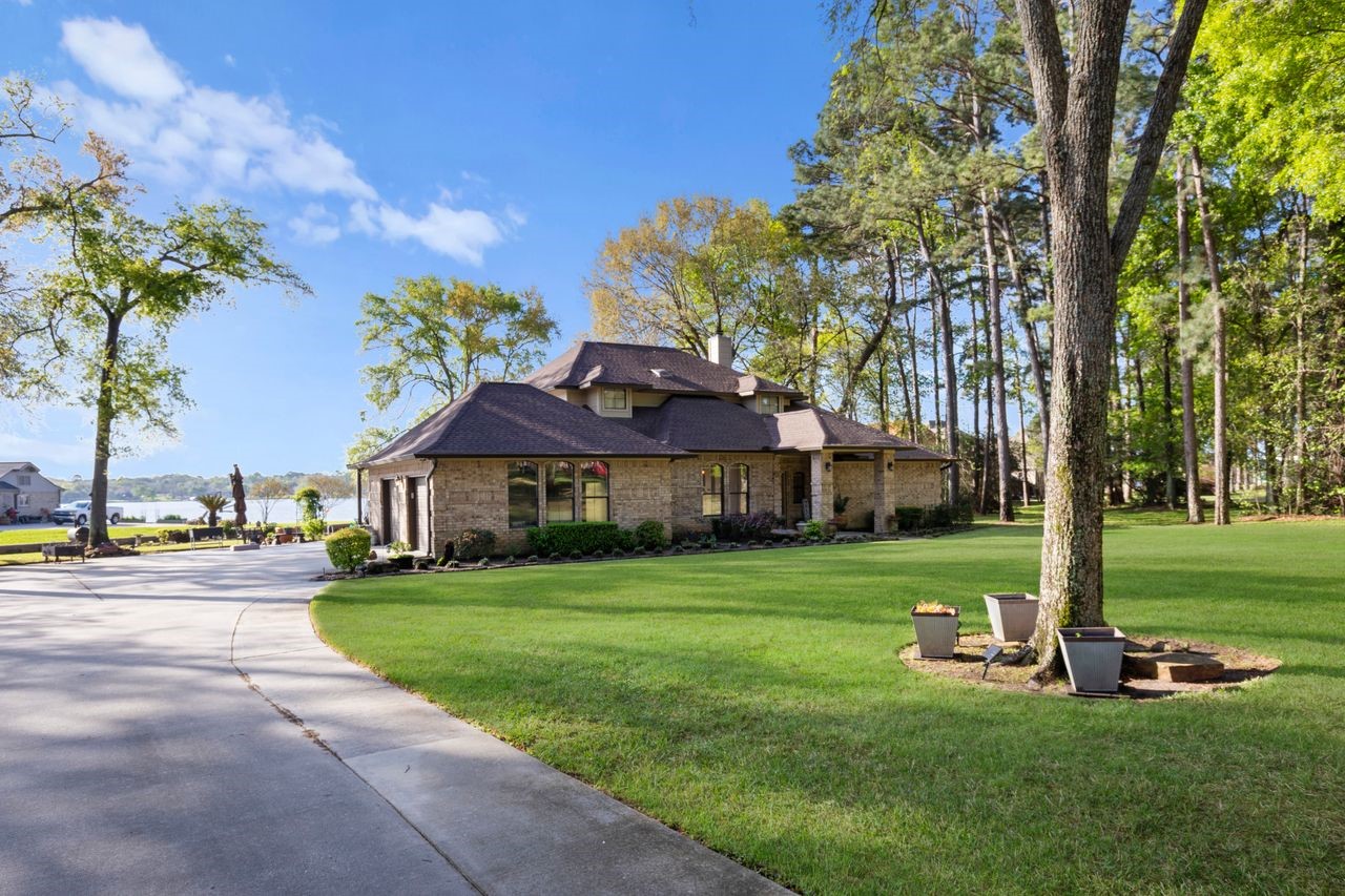 12626 Aries Loop Willis, TX 77318 - Photo 29 of 50 This photo showcases a charming brick home with a well-maintained lawn and a spacious driveway. Surrounded by trees, it offers a serene setting, and there's a view of the water in the background, adding to its appeal.