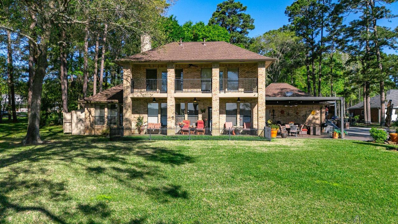 12626 Aries Loop Willis, TX 77318 - Photo 3 of 50 This photo features a charming two-story brick home with a spacious backyard. It has a covered patio area, surrounded by lush greenery and tall trees, providing a serene outdoor space. The house has a classic design with balcony railings and large windows, ideal for enjoying the peaceful surroundings.