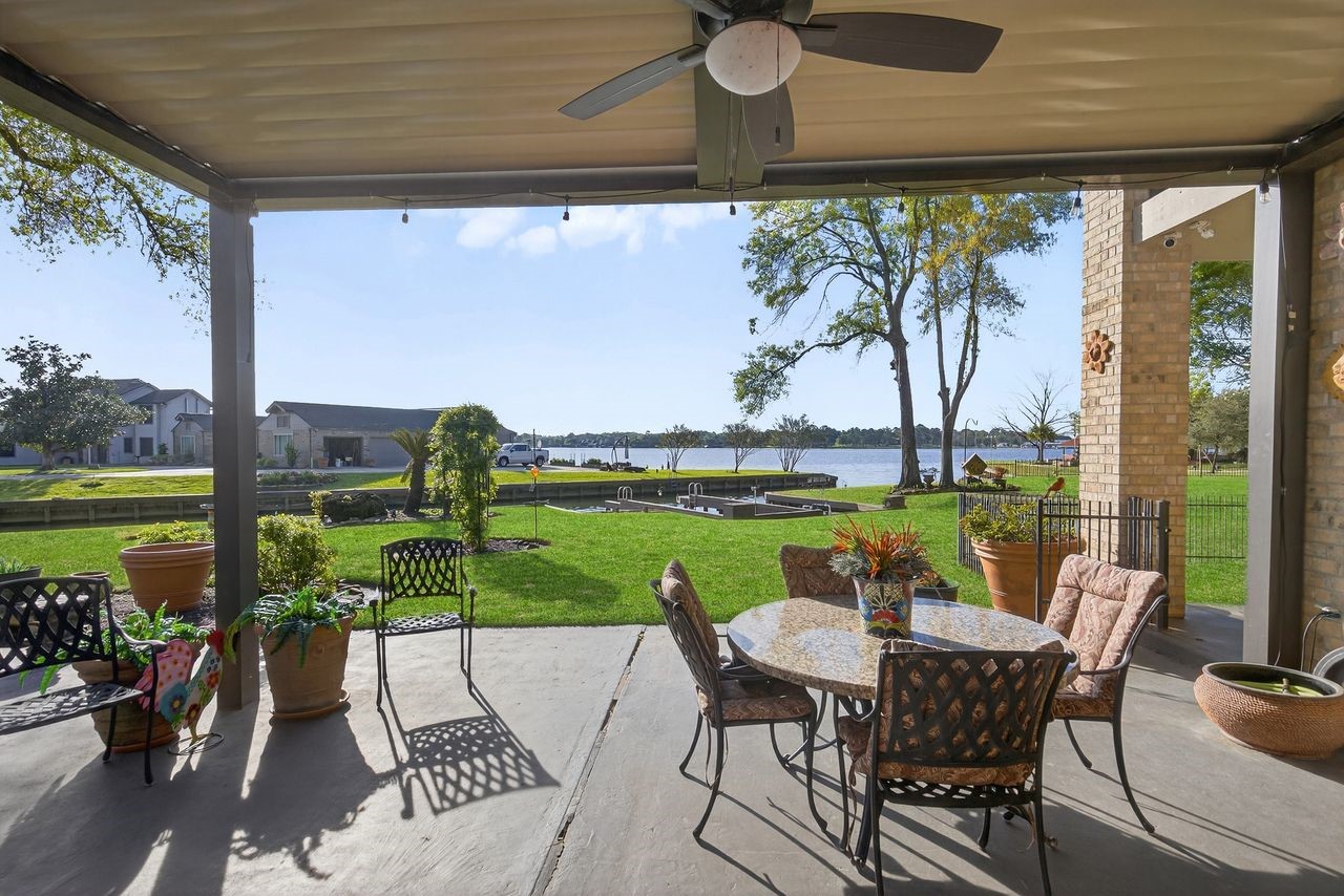 12626 Aries Loop Willis, TX 77318 - Photo 35 of 50 This photo showcases a spacious, covered patio with a ceiling fan, furnished with a table and chairs. It overlooks a lush green lawn, a serene lake, and features a clear, sunny view, ideal for outdoor relaxation and entertaining.