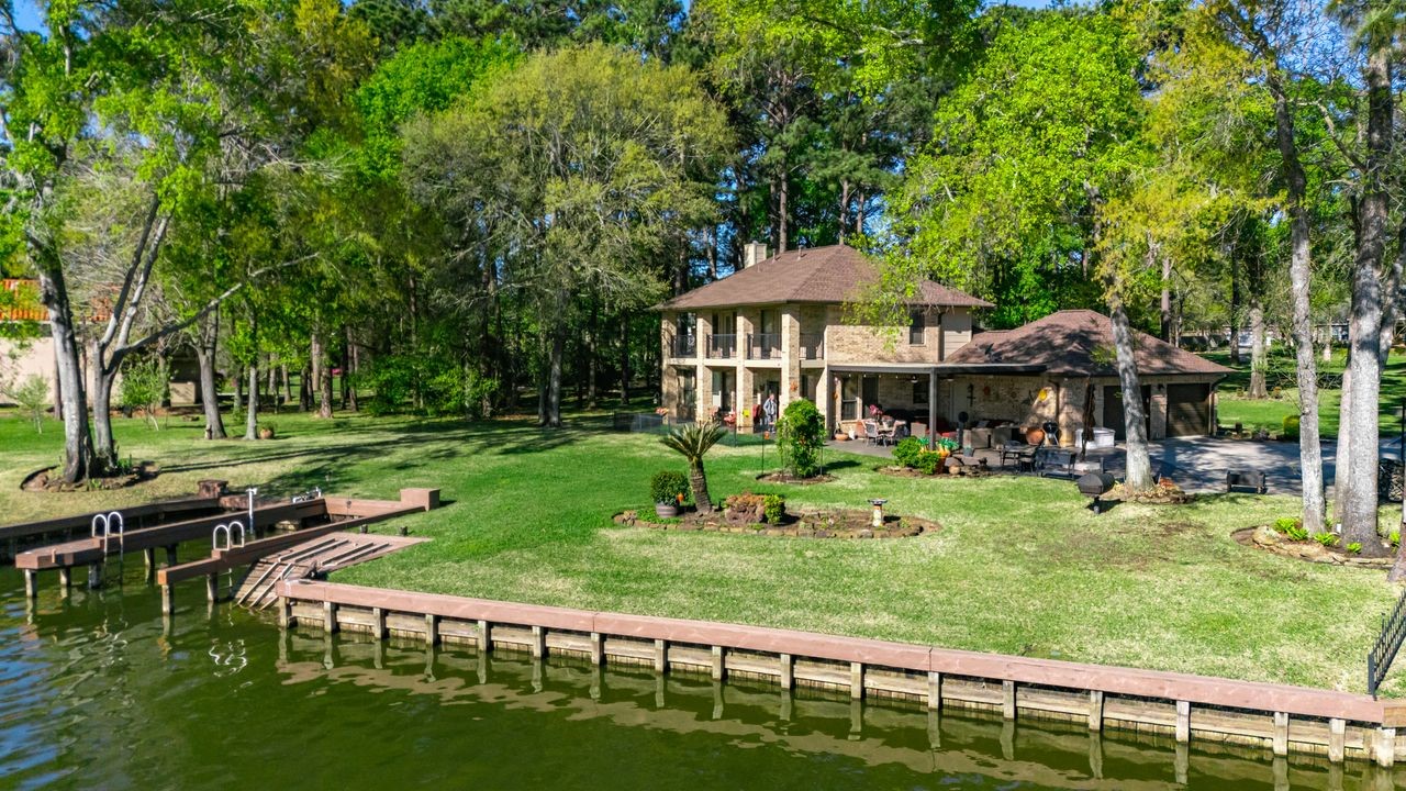 12626 Aries Loop Willis, TX 77318 - Photo 43 of 50 This lakefront property features a charming two-story house surrounded by lush greenery and mature trees. It includes a private dock, expansive lawn, and a spacious patio, perfect for outdoor relaxation and entertaining.