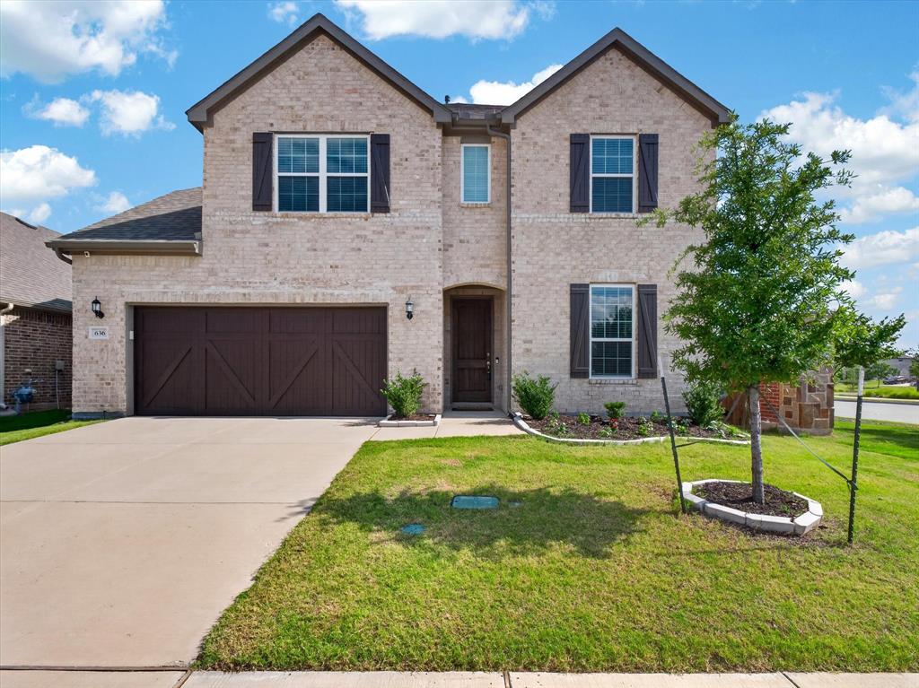 Two-story brick exterior featuring a two-car garage with carriage-style doors, symmetrical gabled roofs, a concrete driveway, and a front yard with a young tree and landscaping