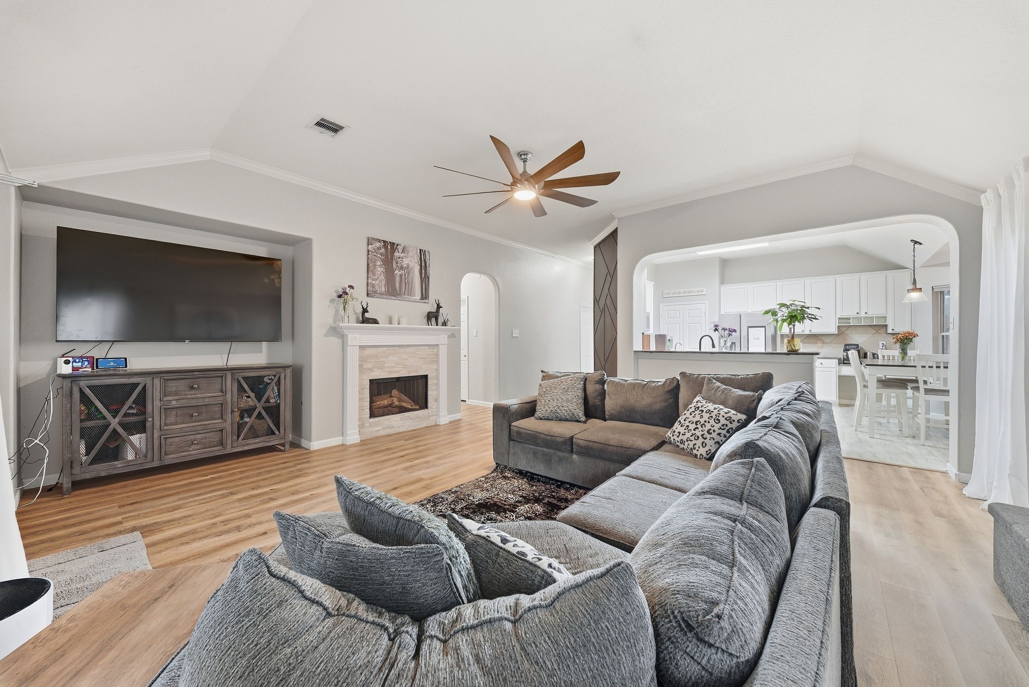 2223 Lazy Trail Path Court Spring, TX 77373 - Photo 8 of 23 Spacious living area with a cozy fireplace, modern ceiling fan, and a large sectional sofa, seamlessly flowing into a bright kitchen with white cabinetry.