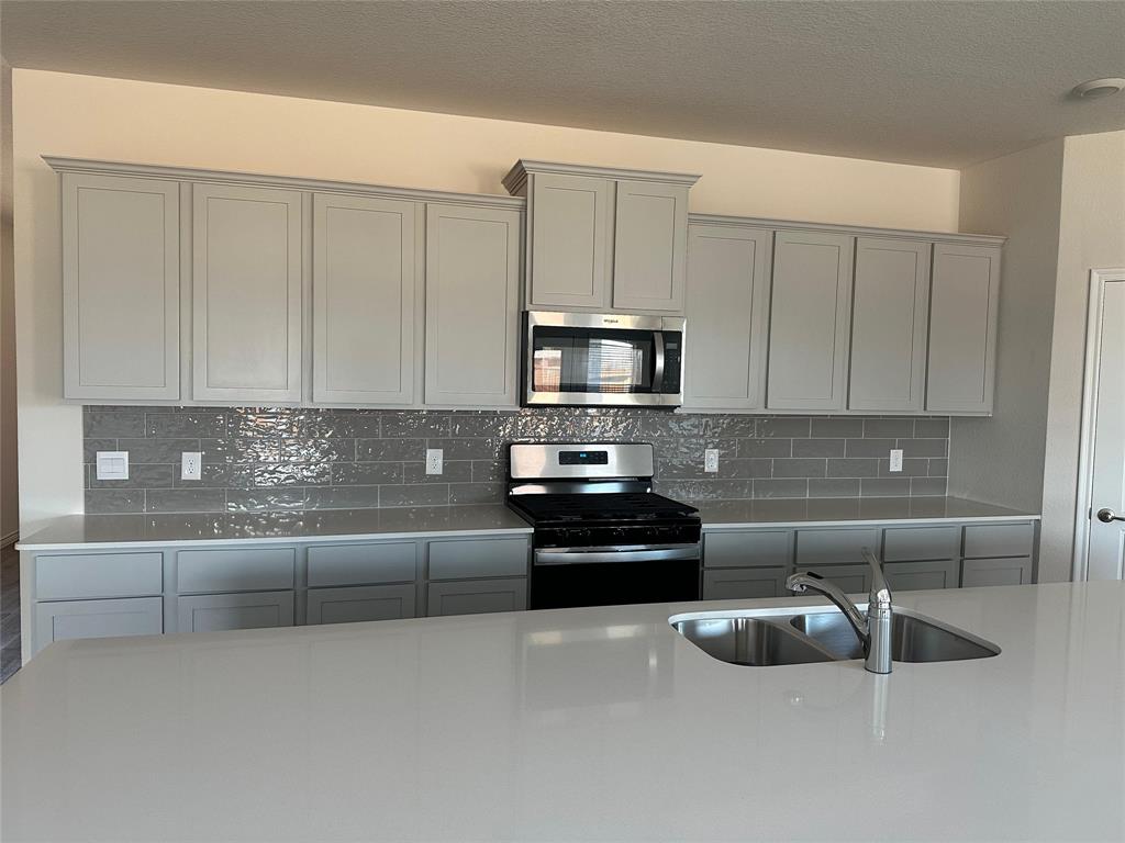 2408 Grapevine Way Celina, TX 75009 - Photo 6 of 20 Kitchen with gray cabinets, stainless steel appliances, backsplash, light stone countertops, and a textured ceiling