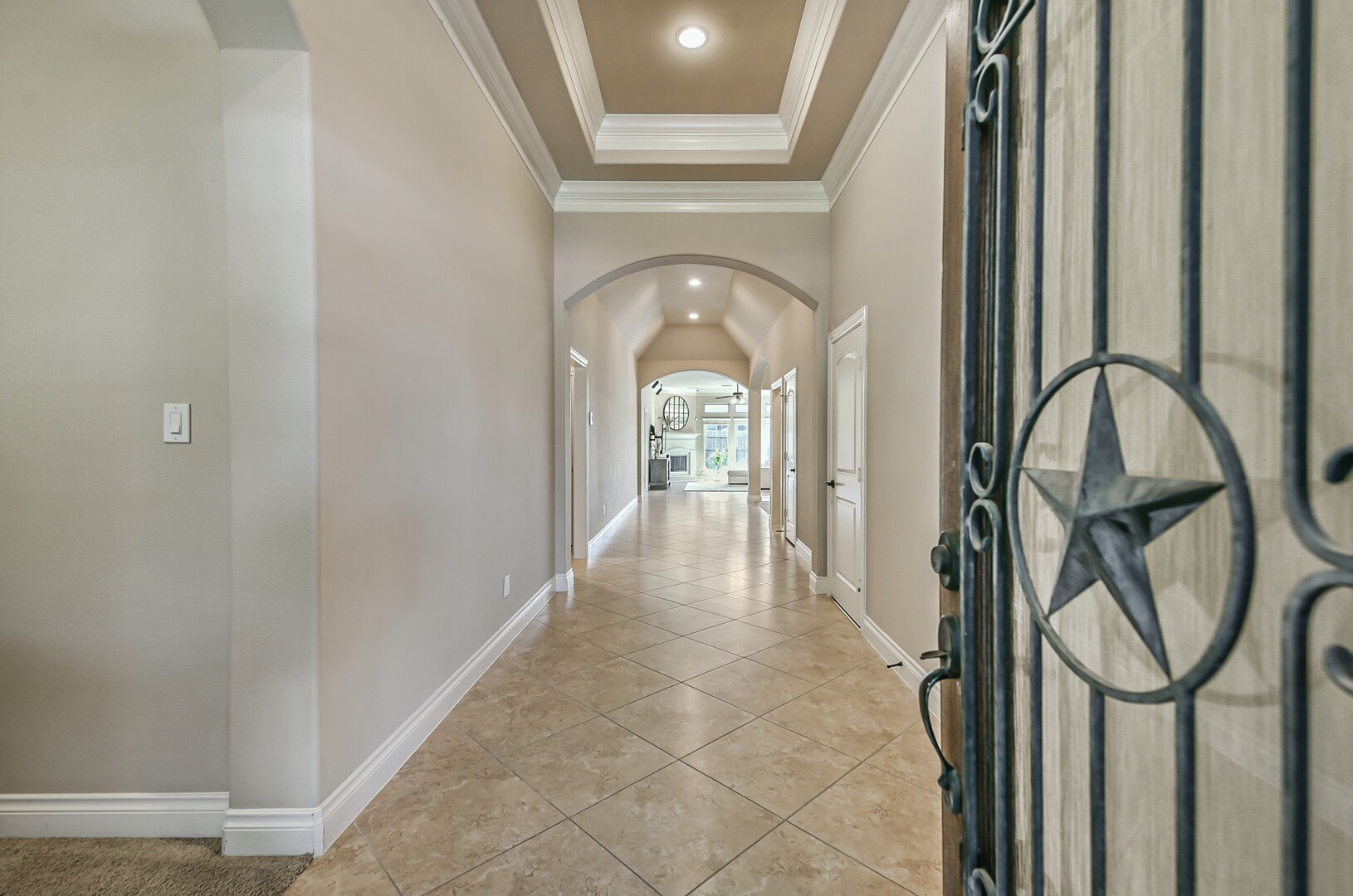 10323 Belvamera Road Richmond, TX 77407 - Photo 4 of 25 This photo showcases a welcoming entrance hallway with elegant tile flooring, soft neutral walls, and a tray ceiling detail. The space is well-lit, with an arched doorway leading to a bright living area in the background.