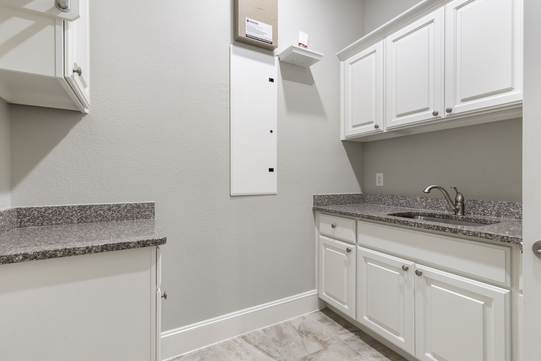 25002 Rosa Aurora Way Spring, TX 77389 - Photo 22 of 38 This photo shows a modern laundry room with light gray walls and tiled flooring. It features white cabinetry, granite countertops, and a stainless steel sink, providing ample storage and workspace.