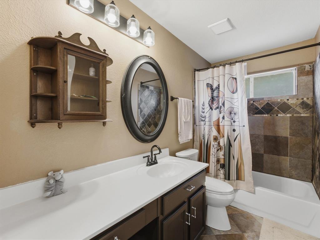 4008 Falcon Ridge Drive Haltom City, TX 76137 - Photo 32 of 40 Bathroom featuring an oval mirror, a four-light vanity fixture, a dark wood-finish vanity with a white countertop and integrated sink, a built-in bathtub with a tiled surround, and multi-toned floor tiling