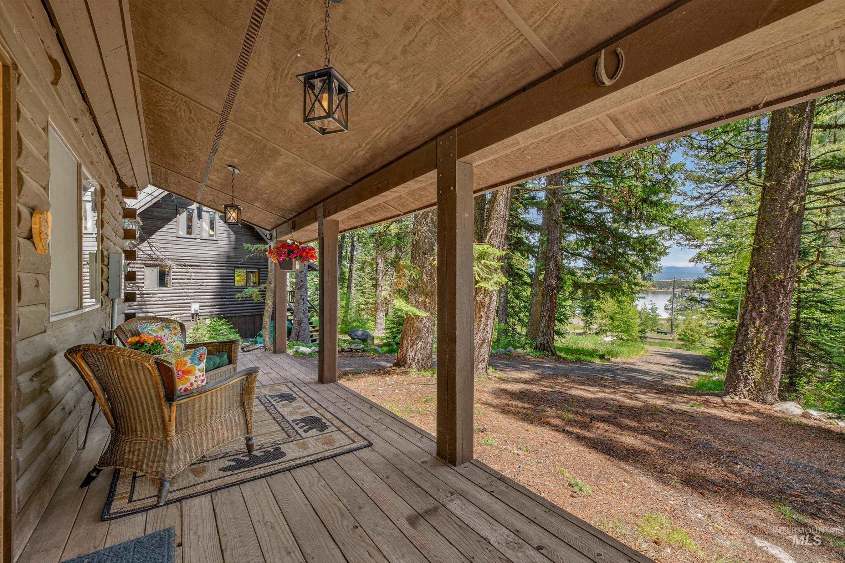 2339 West Mountain Road Donnelly, ID 83615 - Photo 28 of 37 Covered wood-plank porch with exposed wood beams, featuring two black hanging lanterns and a natural wooded backdrop with distant water
