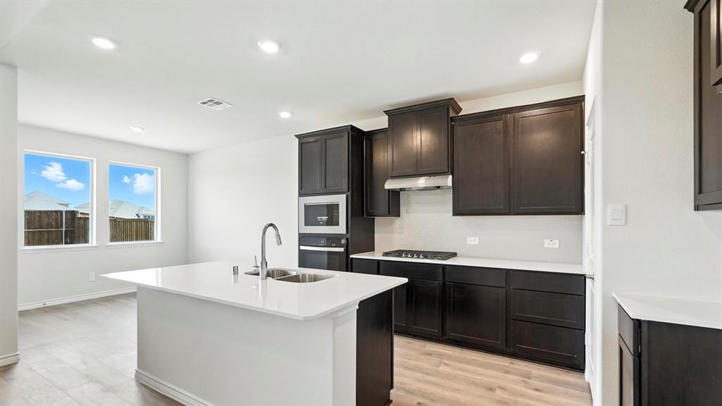 4159 Waldron Trail Forney, TX 75126 - Photo 3 of 19 The kitchen features an island with a dual basin sink, dark wood cabinetry, white countertops, a built-in microwave, and a gas cooktop with an overhead range hood
