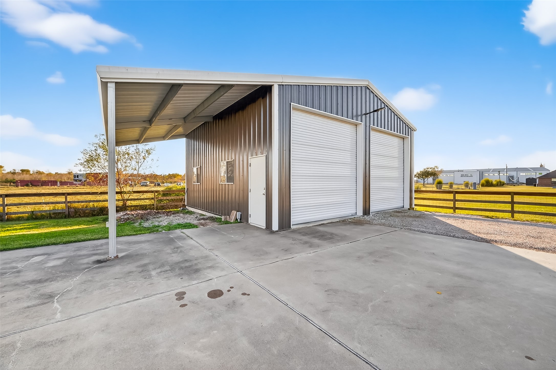 912 County Road 340 Angleton, TX 77515 - Photo 33 of 50 This photo shows a spacious metal building with two large roll-up doors, ideal for storage or workshop use. It features a covered area on one side and is set on a concrete driveway with rural surroundings, offering ample space and versatility.