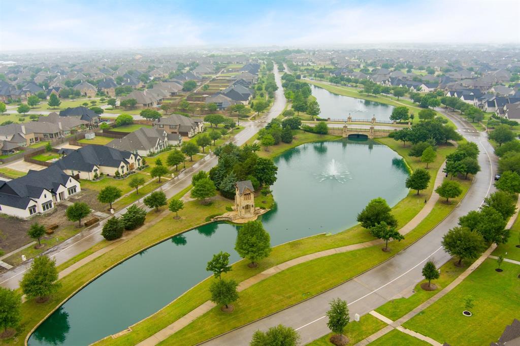 1501 Calcot Lane Forney, TX 75126 - Photo 33 of 34 Expansive residential area featuring a central waterway with a fountain, a decorative pedestrian bridge, lush green landscaping, a stone-clad tower structure, and numerous homes with dark-pitched roofs