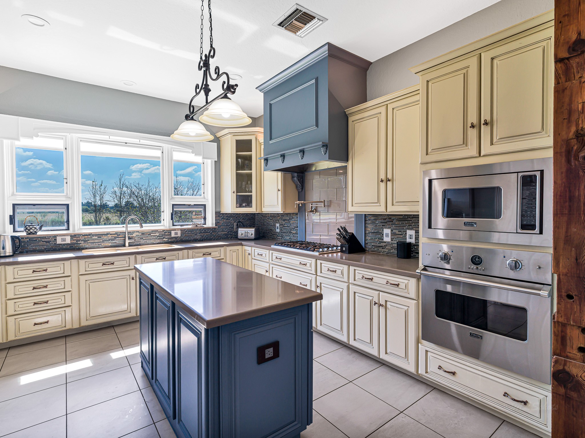 100 Pecan Grove Road Sealy, TX 77474 - Photo 16 of 48 This photo showcases a bright, spacious kitchen with a large window providing natural light. The kitchen features elegant cabinetry, a central island with a blue finish, stainless steel appliances, and a stylish light fixture. The open wooden doorway leads to a cozy adjoining room, adding warmth and character.