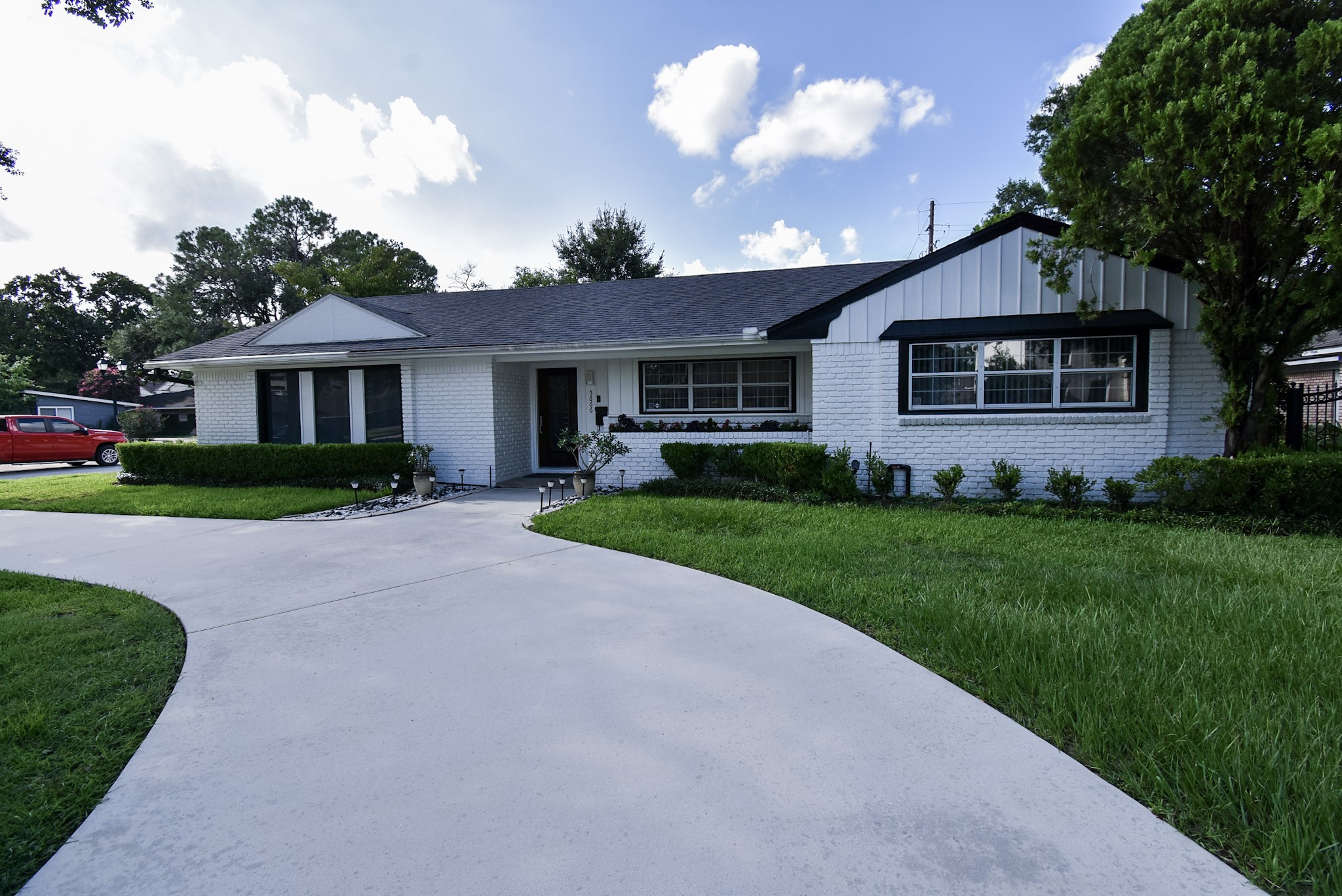 Charming single-story white brick home with large front windows and a neatly manicured lawn. It has a curved driveway and is surrounded by well-maintained shrubs and trees, offering great curb appeal.