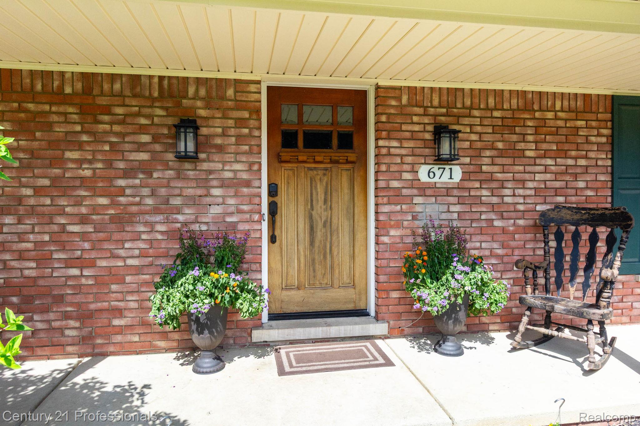 671 Bridgestone Drive Rochester, MI 48309 - Photo 4 of 50 Brick exterior featuring a paneled wood-finish entry door with clear glass accents, twin wall-mounted lanterns, and a concrete porch
