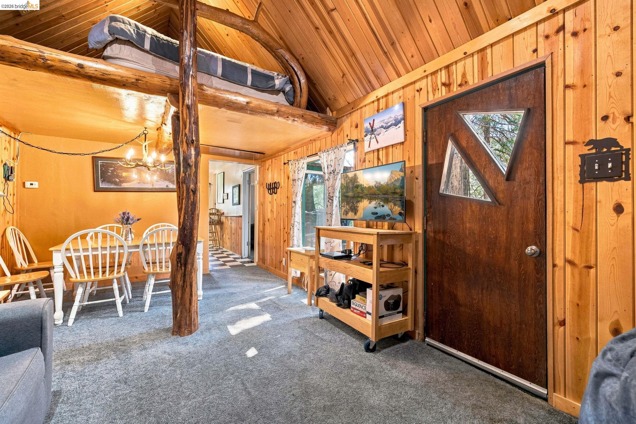 26457 Crystal Road Long Barn, CA 95335 - Photo 2 of 39 Rustic wood-paneled interior featuring a lofted sleeping area, exposed log support beams, a dining area with pendant lighting, and a unique entry door with triangular window inserts