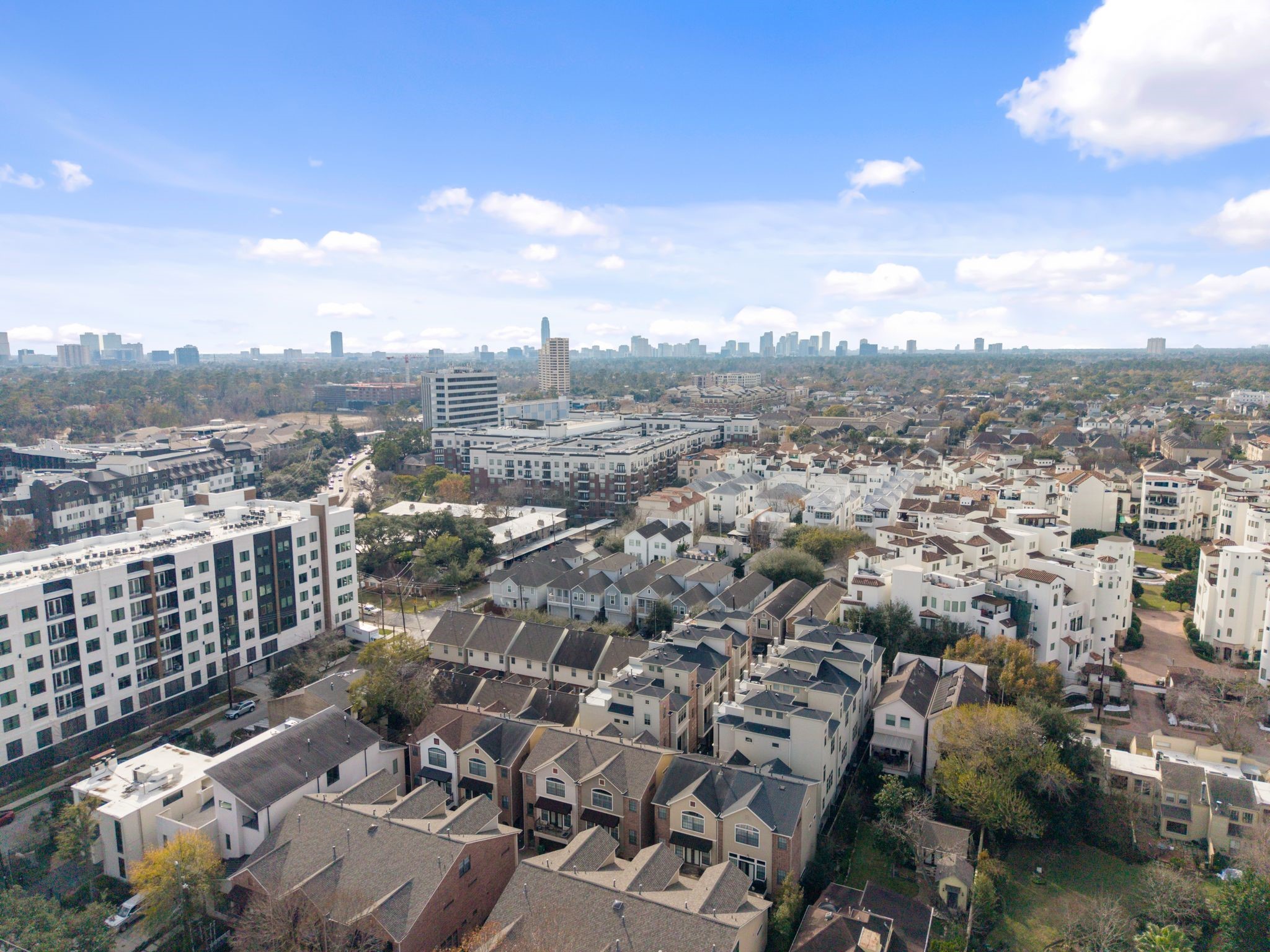 121 Roy Street Houston, TX 77007 - Photo 26 of 29 Aerial view of a suburban neighborhood featuring a mix of modern townhouses and mid-rise apartment buildings. The area is lush with greenery and offers a glimpse of the city skyline under a bright blue sky.