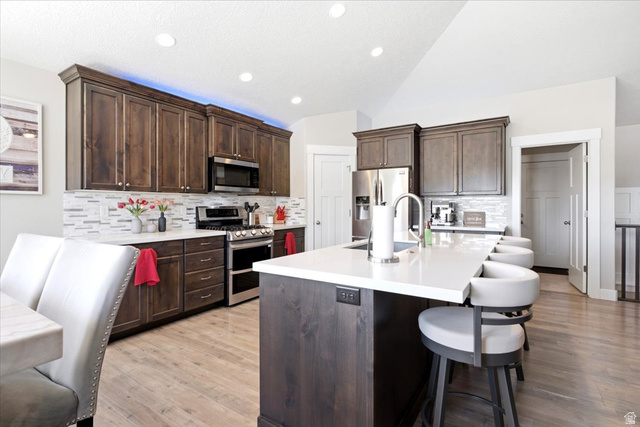 2973 West Red Sand Rd Haven Ogden, UT 84401 - Photo 10 of 30 Spacious kitchen featuring dark wood-finish cabinetry, stainless steel appliances, a white tile backsplash, a large kitchen island with seating, and light wood-finish flooring