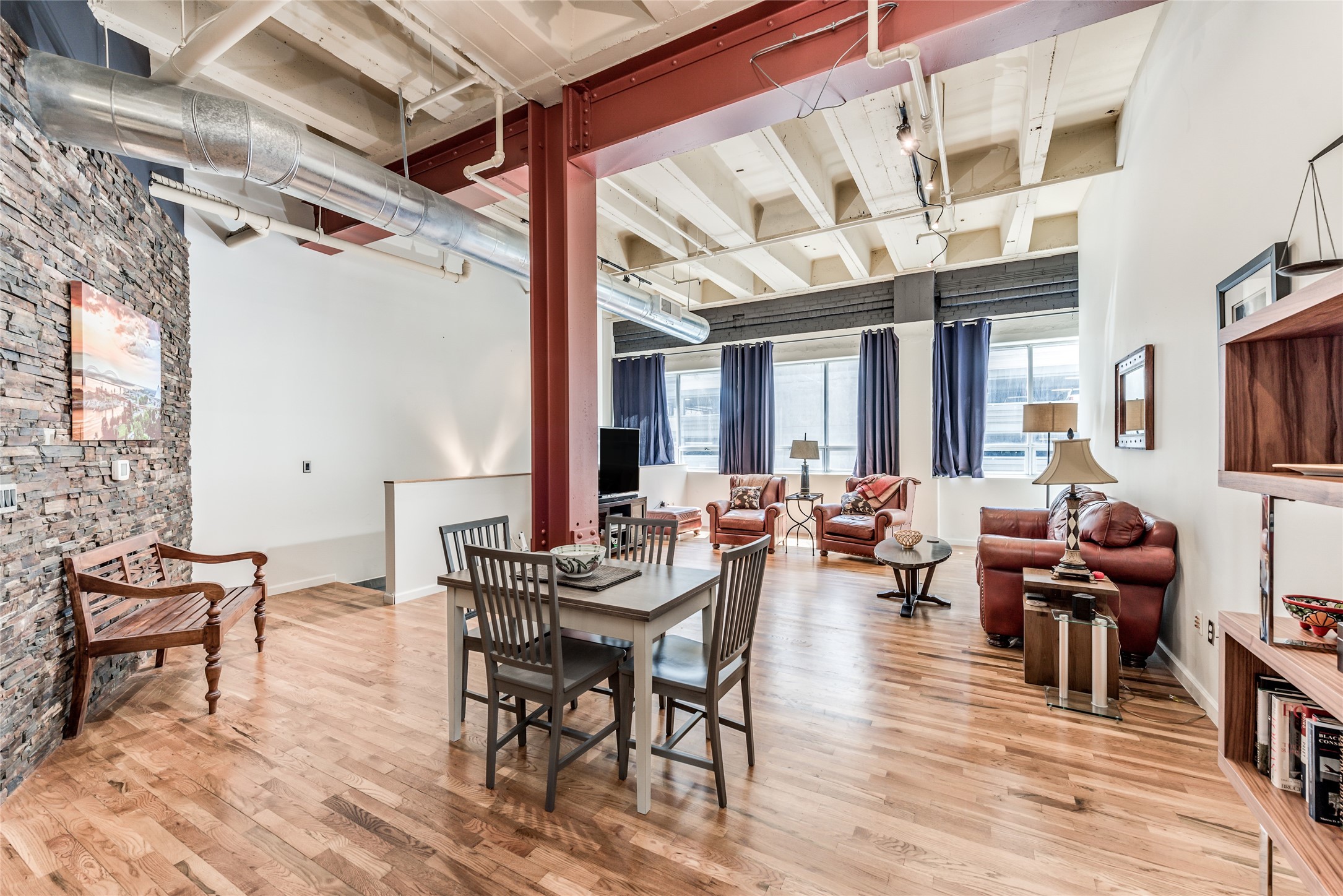 705 Main Street, Unit 215 Houston, TX 77002 - Photo 6 of 36 This photo showcases a spacious, open-concept loft with high ceilings, exposed ductwork, and a mix of industrial and cozy elements. The room features polished wood floors, a brick accent wall, and large windows with ample natural light. The living area includes comfortable seating and a dining space, perfect for modern urban living.