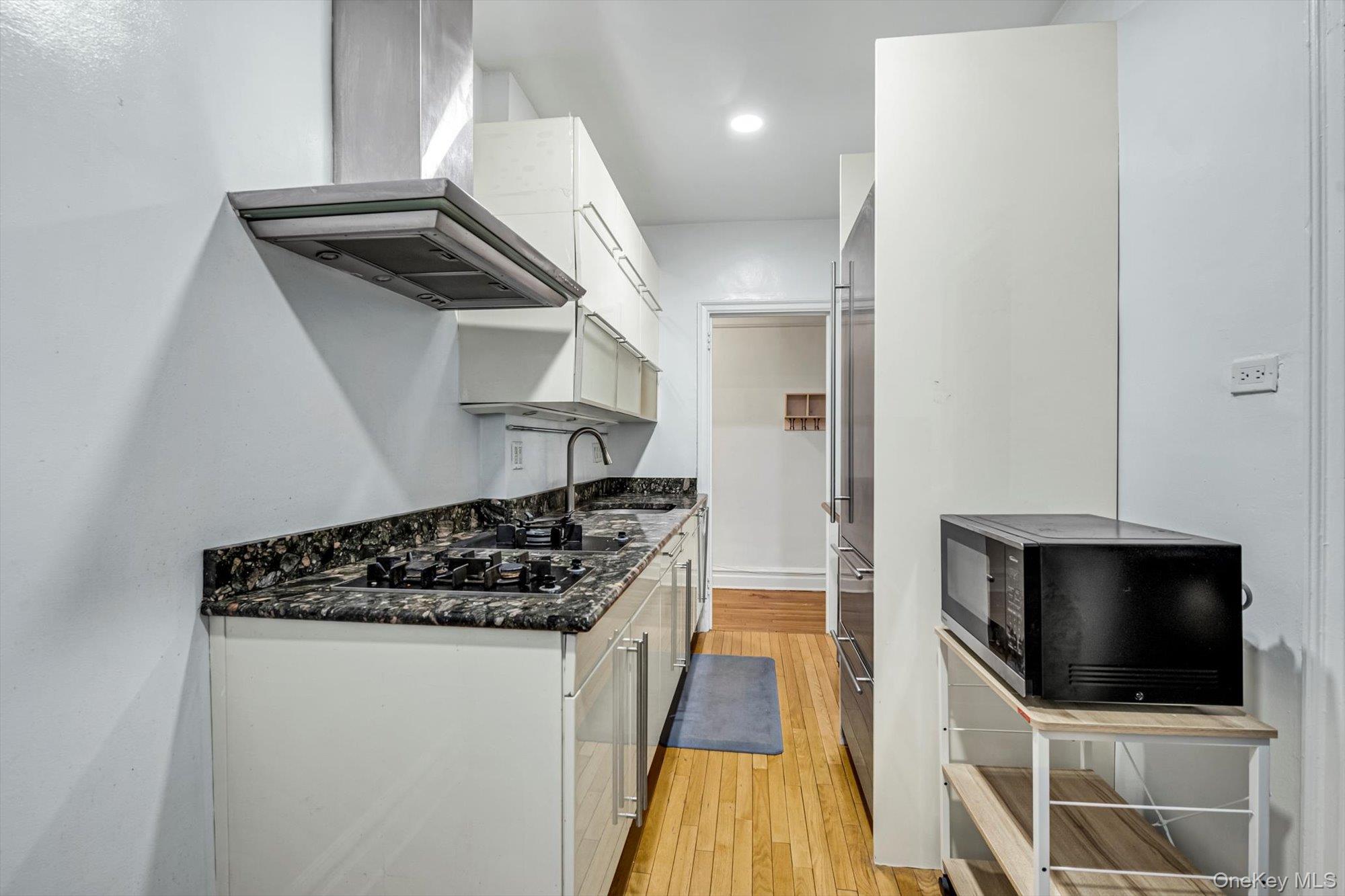 72-34 Austin Street, Unit F10 Queens, NY 11375 - Photo 7 of 15 Kitchen with white cabinets, wall chimney range hood, black microwave, dark stone counters, and light wood-type flooring