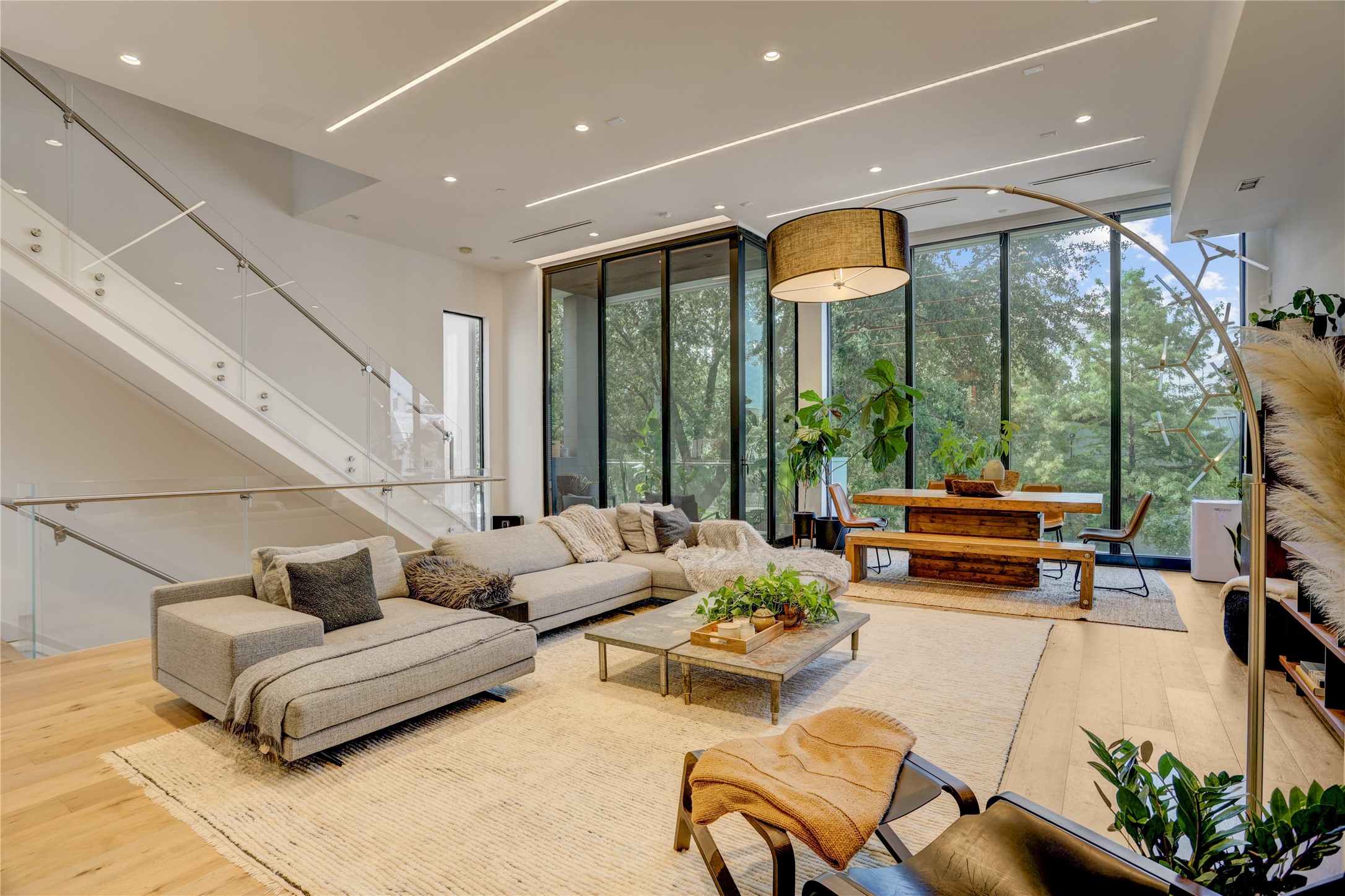 1807 Indiana Street Houston, TX 77006 - Photo 7 of 35 Living & Dining Area: Bright, open living space framed by a modern glass-panel staircase, featuring architectural linear lighting, wide-plank wood flooring, and floor-to-ceiling windows looking out to lush treetop views.