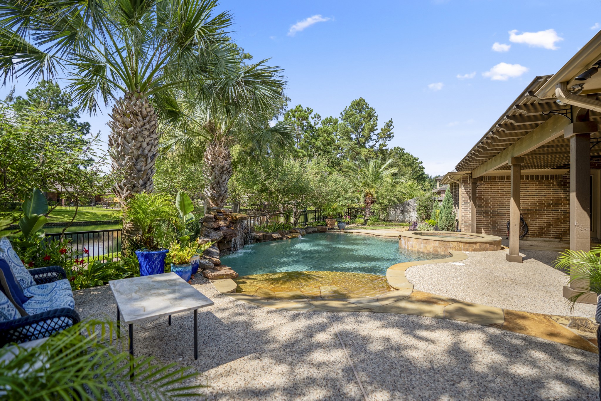 2407 Hendricks Lakes Drive Spring, TX 77388 - Photo 39 of 50 Poolside from the far end of the backyard, this view captures the beach-style pool entry, raised spa, and water beyond the yard for a fuller sense of the outdoor setting. The covered pergola stretches along the right side of the home, while palm trees and layered landscaping wrap the space with a relaxed, resort-style feel made for slow evenings, weekend swims, and time outside.