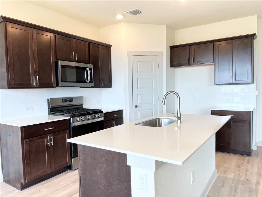10600 Defender Trail Austin, TX 78754 - Photo 6 of 21 Kitchen with dark wood finish cabinetry, stainless steel appliances, tasteful backsplash, a center island with sink, and light wood-style floors