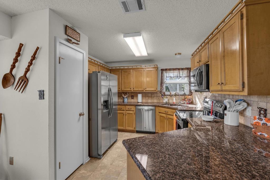107 Fort Graham Circle Waco, TX 76705 - Photo 10 of 40 Kitchen with appliances with stainless steel finishes, brown cabinetry, decorative backsplash, dark stone counters, and a textured ceiling