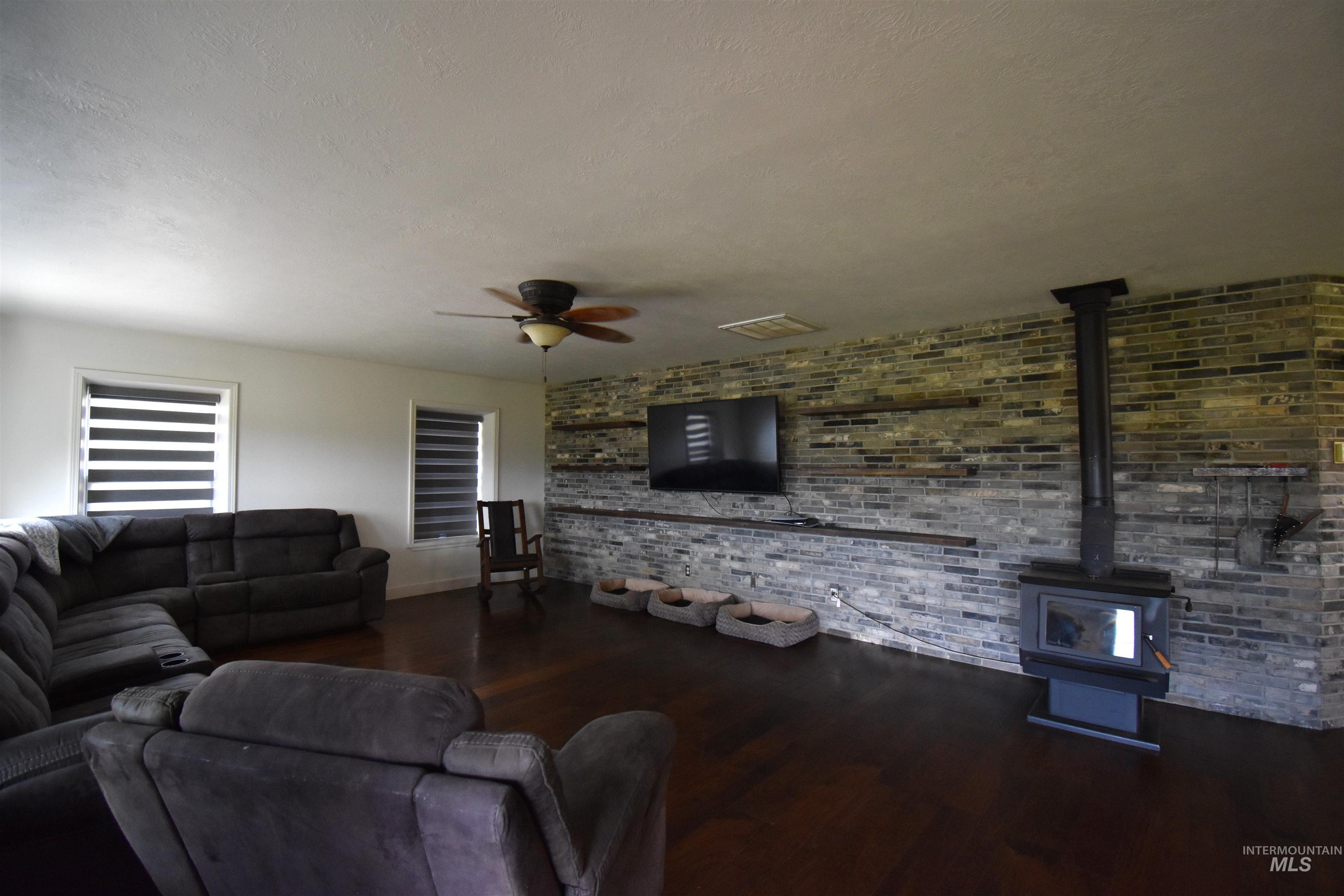 2599 Fruitvale Glendale Road Council, ID 83612 - Photo 9 of 38 Living room featuring dark floors, a ceiling fan, a wood stove, and a brick accent wall.